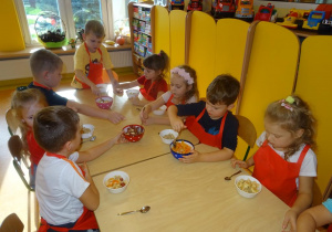 Grupa dzieci siedzi przy stole i nakłada łyżkami owoce do swoich misek.